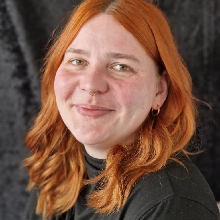 Laura Sandberg pHKP Lächelnde Person mit roten Haaren und schwarzem Oberteil vor schwarzem Hintergrund.