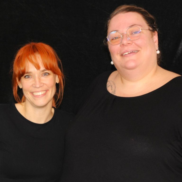 Inke Peters und Ulrike Kloninger | Geschäftsführerinnen Zwei gestandene Frauen lächeln vor einem dunklen Hintergrund.